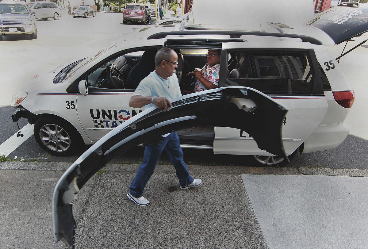Manuel Abreu, a manager at Union Taxi, carries a bumper that was ripped off the taxi that was involved in an accident on Lewis Street. Lauren Snow of Lynn is sitting in the taxi.
